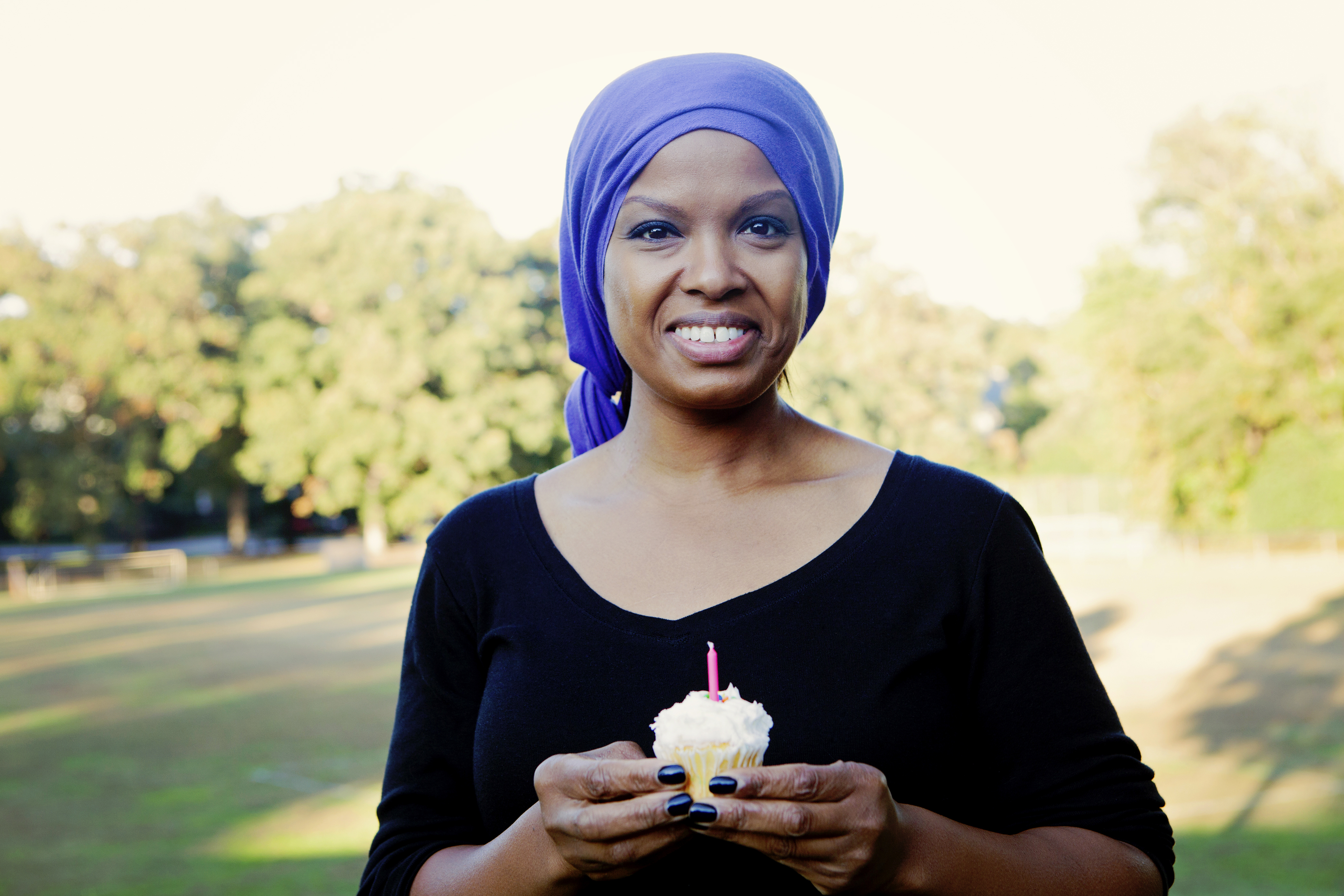 A woman with cancer wearing a scarf on her head and holding a cupcake with a pink candle