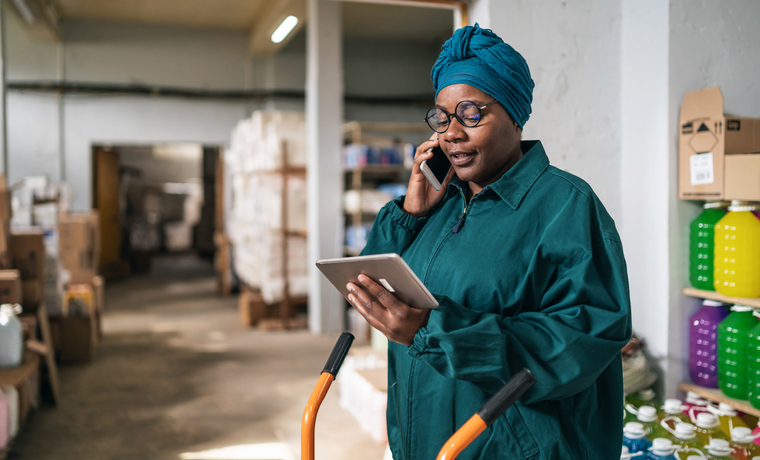 Afro female warehouse manager using a hand truck and talking on a smart phone in distribution warehouse