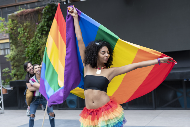 Portrait of a brunette Latina with curly hair enjoying her gay pride day dressed very colorful smiling at the camera while happily holding her gay pride flag