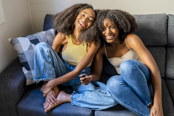 Young woman with remote control laughing by girlfriend in living room - holiday gift guide