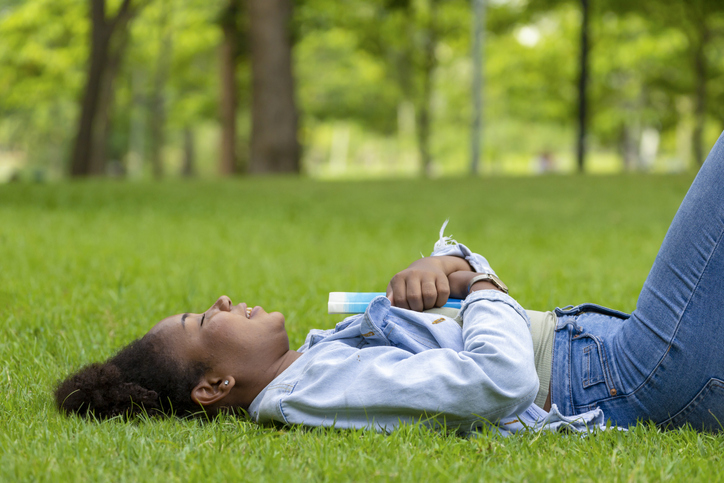 African American woman is lying down in the grass lawn inside the public park holding book in her hand during summer for reading and education concept