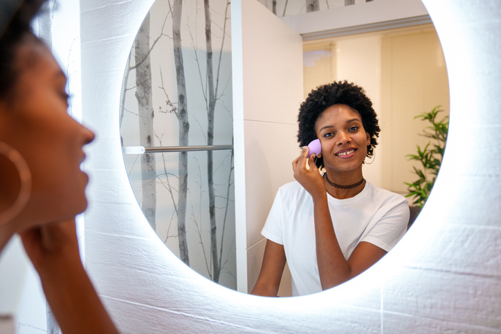 Afro woman happily applying makeup in front of a bright round mirror in a modern bathroom.