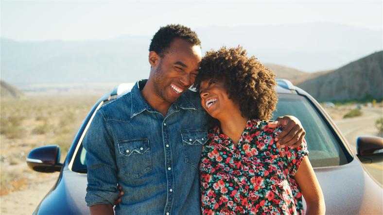 Smiling Young Couple On Road Trip Standing By Rental Car On Country Road