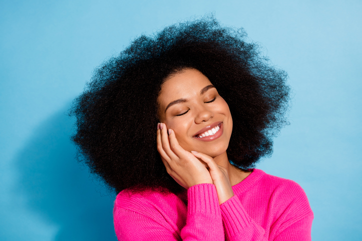 Smiling young woman against a blue background wearing a pink sweater, enjoying a relaxed moment, with afro hairstyle