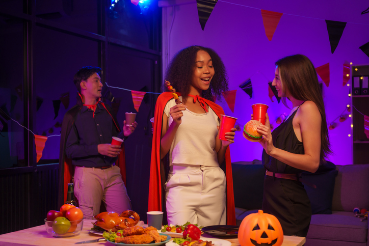 An African American woman and a white woman celebrate with coworkers, drinking, having fun, and smiling together at a lively Halloween night party filled with joy and laughter.
